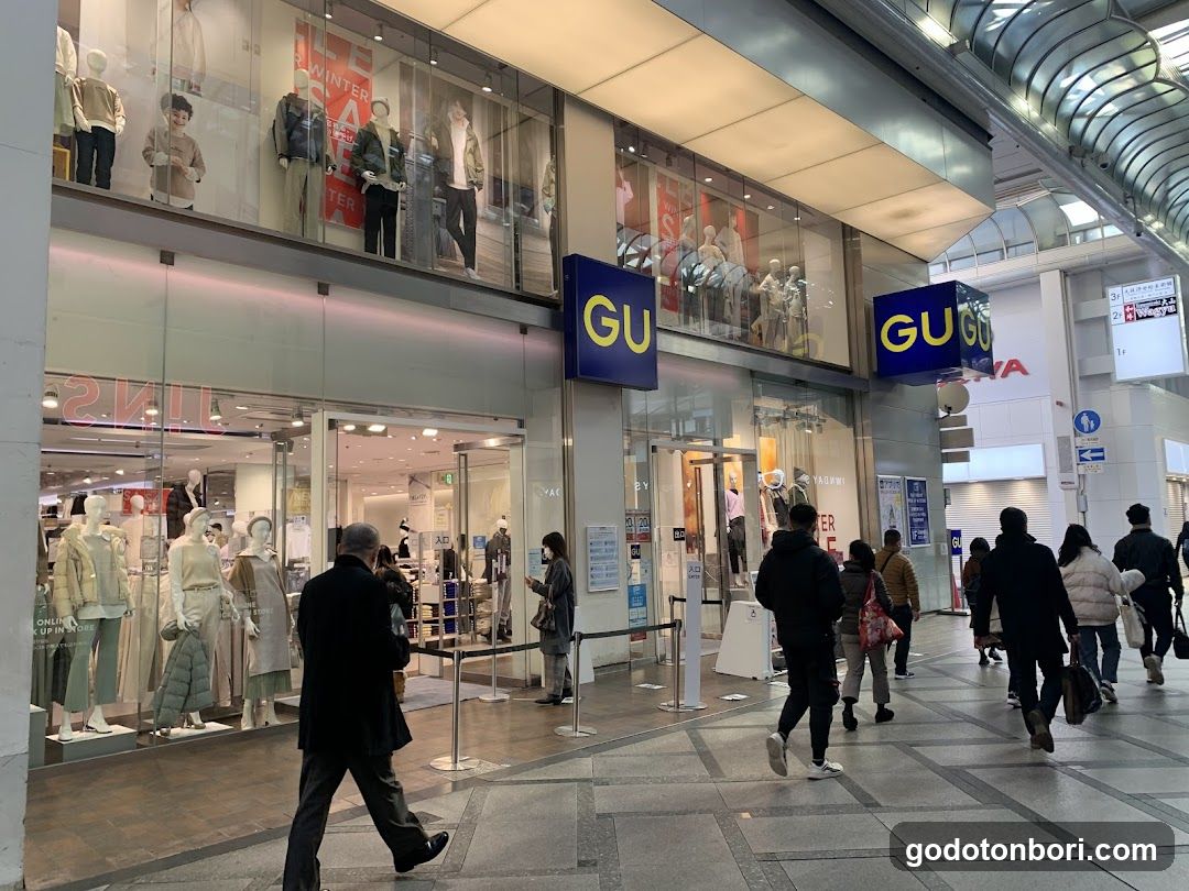 A modern retail storefront with large glass windows displaying mannequins in winter clothing. The GU logo is prominently displayed on blue signs above the entrance. Pedestrians walk along a tiled corridor outside, and an escalator is visible in the background.