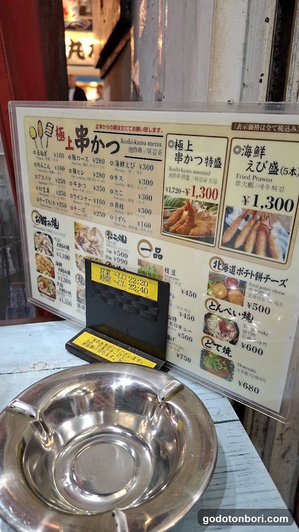 A menu board displaying various Koshikatsu dishes with prices in yen, including items like 'Koshikatsu menu', 'Fried Prawns', and 'Hokkaido Potato Cheese'. The menu is placed on a table next to an ashtray.