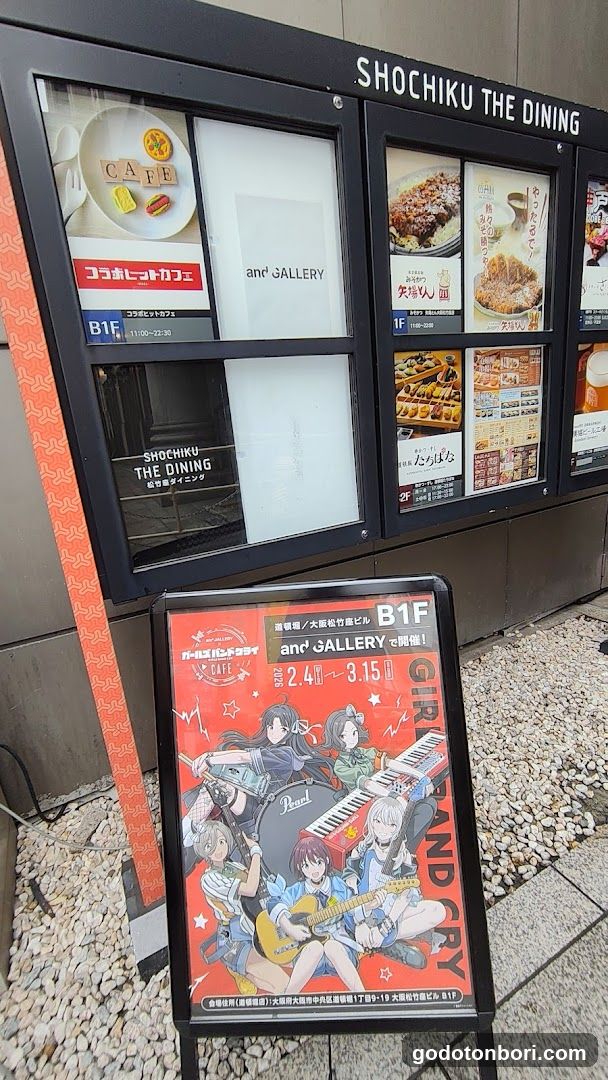 Exterior view of Shochiku The Dining storefront with visible signage on the building facade. A menu board displays food items including 'Kokobito Coffee', 'and GALLERY', and various dishes like 'Takoyaki' and 'Tofu'. An anime-themed poster advertises an event from April 24 to May 31.