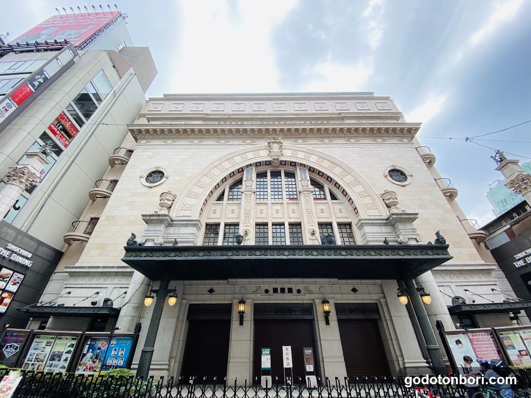 A grand, classical-style building with a large arched entrance and decorative stonework. Visible signage includes 'SUSHI NO THE DINING' on the left side of the facade. The main entrance features dark wooden doors under a green awning with illuminated lamps.