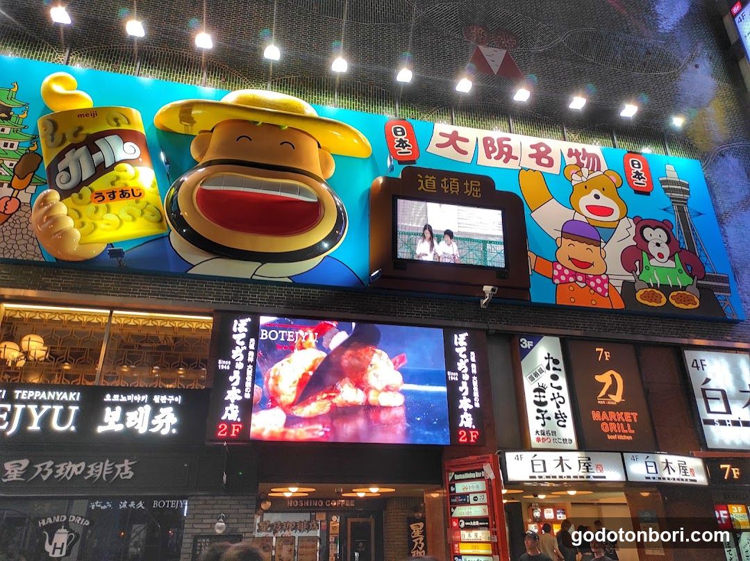 A vibrant storefront featuring a large cartoon character holding a cup of udon, with multiple shop signs below including 'BOTEIYU', 'TEPPANYAKI', and 'MARKET GRILL'. The building is illuminated at night with bright signage.
