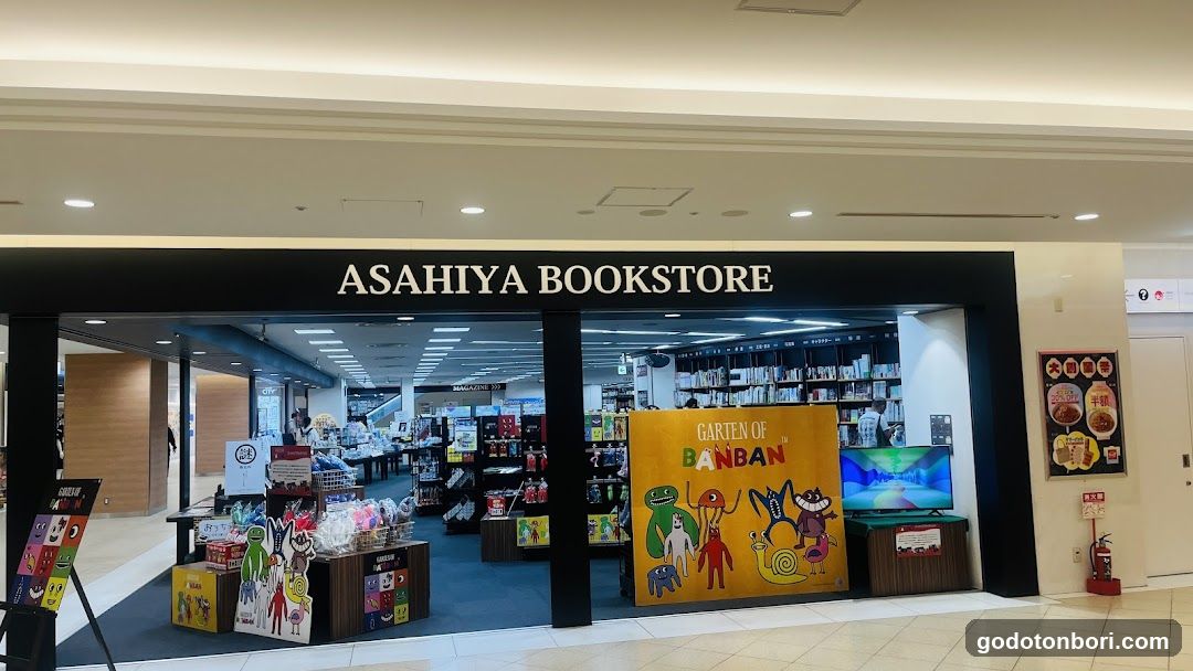 The image shows the storefront of Asahiya Bookstore, a bookstore located in a shopping mall or similar indoor space. The store's name is prominently displayed on a black sign above the entrance. Inside, shelves filled with books are visible, along with various promotional displays and merchandise. A large poster featuring characters from 'Garten of Banban' is placed near the entrance, adding to the colorful and inviting atmosphere.