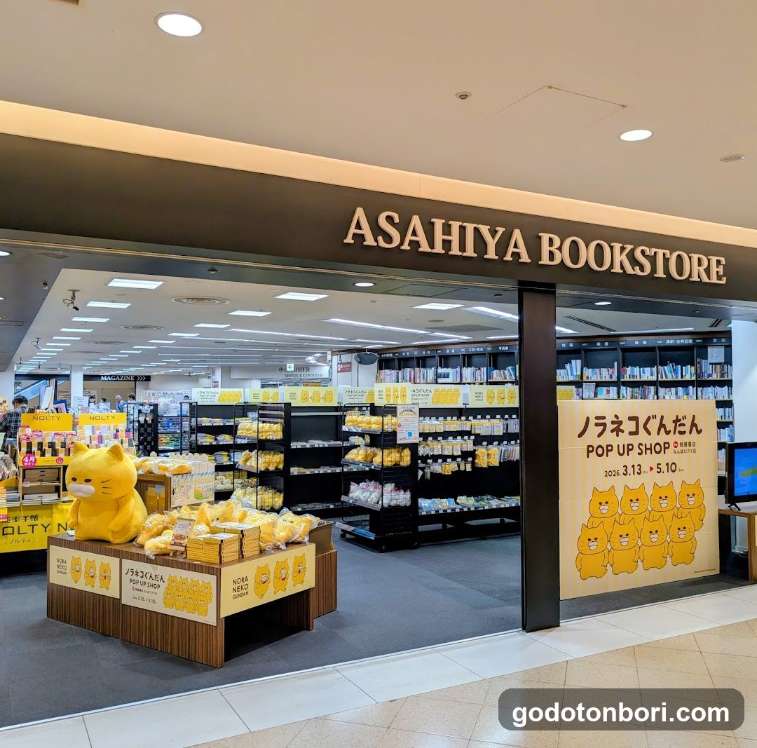 The image shows the storefront of Asahiya Bookstore, featuring a prominent sign above the entrance. Inside, shelves are stocked with books and merchandise. A large yellow cat plush toy sits on a display table in front of the store. To the right, there is a poster advertising a 'Pop Up Shop' event.