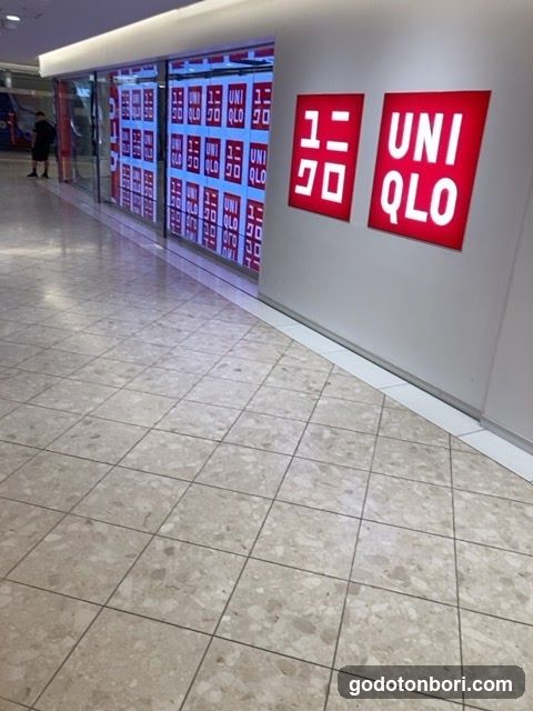 Photo of a Uniqlo storefront with large red signage displaying the brand name and logo on the wall. The entrance features glass doors covered in promotional posters with repeated 'UNIQLO' branding.