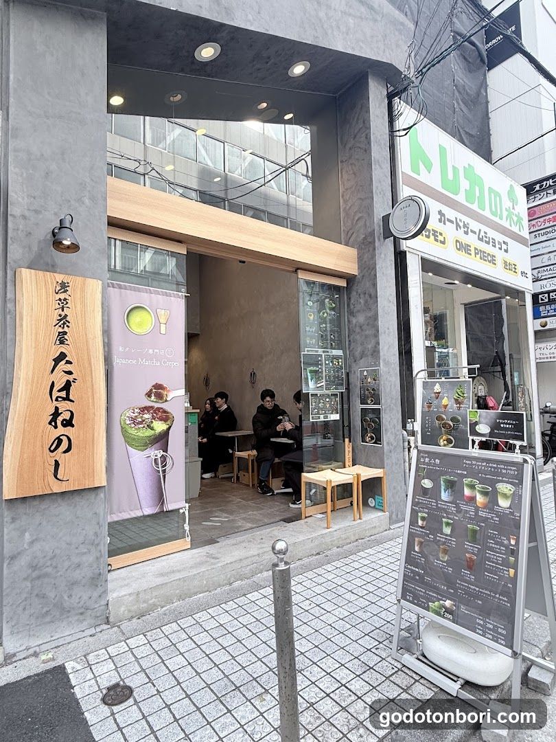 The image shows the exterior of a shop with two distinct entrances. On the left, there is a wooden sign reading 'Ihane Matcha Crisp' (淡葉茶屋たばねのし) above an open doorway leading into a cafe-like space where customers are seated at tables. A large poster advertising matcha drinks hangs near the entrance. To the right, another storefront features signage for 'Toreka no Soba' and mentions 'ONE PIECE', indicating it may be a card game or anime-themed shop. Outside, a menu board displays various drink options with prices.